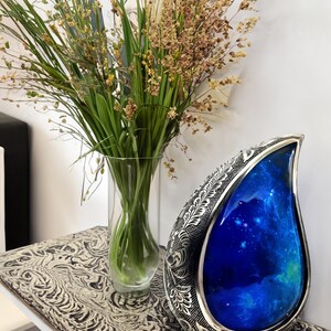 Op de afbeelding: Een zilveren urn in de vorm van een traan met een blauw galaxy-ontwerp. De urn staat op een tafel met een gepatroneerd oppervlak. Op de achtergrond staat een vaas met groene planten.