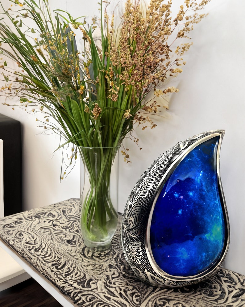 Op de afbeelding: Een zilveren urn in de vorm van een traan met een blauw galaxy-ontwerp. De urn staat op een tafel met een gepatroneerd oppervlak. Op de achtergrond staat een vaas met groene planten.