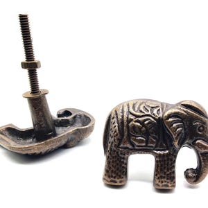 May include: Two antique bronze elephant-shaped cabinet knobs. One knob is fully assembled with a screw and nut, while the other is a decorative elephant. The elephant knob has detailed carvings and a vintage finish.