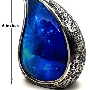Op de afbeelding: Een zilveren urn in de vorm van een traan met een gegraveerd bloemenmotief. De urn heeft een blauw, galaxy-thema interieur en is 15,2 cm hoog en 11,4 cm breed.