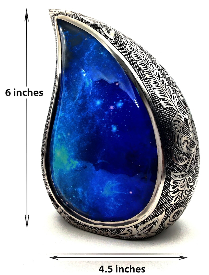 Op de afbeelding: Een zilveren urn in de vorm van een traan met een gegraveerd bloemenmotief. De urn heeft een blauw, galaxy-thema interieur en is 15,2 cm hoog en 11,4 cm breed.