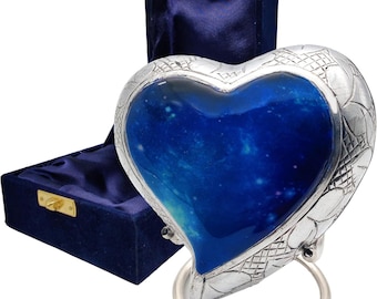 Puede incluir: Urna en forma de corazón con un diseño de galaxia azul y blanco, enmarcada en un marco de metal plateado. La urna tiene un anillo plateado en la parte inferior y se muestra junto a una caja de terciopelo azul.