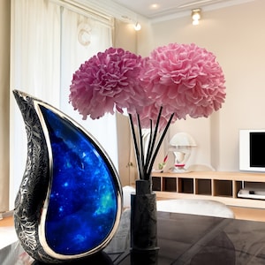 Op de afbeelding: Een zilveren urn in de vorm van een traan met een blauw galaxy-ontwerp staat op een zwarte tafel naast een vaas met roze bloemen.