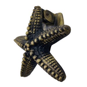 May include: A bronze-colored starfish-shaped cabinet pull. The five-pointed star has a textured surface with small, round details. The hardware is attached to the back for easy installation. Ideal for coastal or nautical decor.
