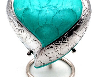 Puede incluir: Una urna de corazón de plata con un acabado de esmalte azul turquesa. La urna está sobre un soporte de plata y tiene un diseño floral detallado.
