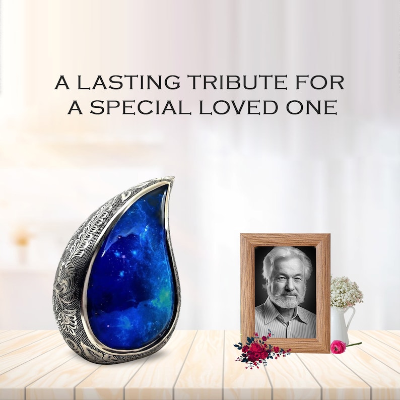 Op de afbeelding: Een zilveren urn in de vorm van een traan met een blauw galaxy-ontwerp. De urn staat op een houten oppervlak naast een ingelijste foto van een man met een witte baard.