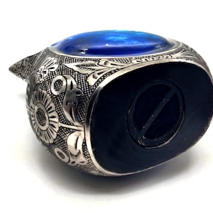 Op de afbeelding: Een zilveren metalen spaarpot in de vorm van een vogel met een blauw glazen oog. De spaarpot heeft een ingewikkeld bloemmotief en een gleuf aan de onderkant voor munten.