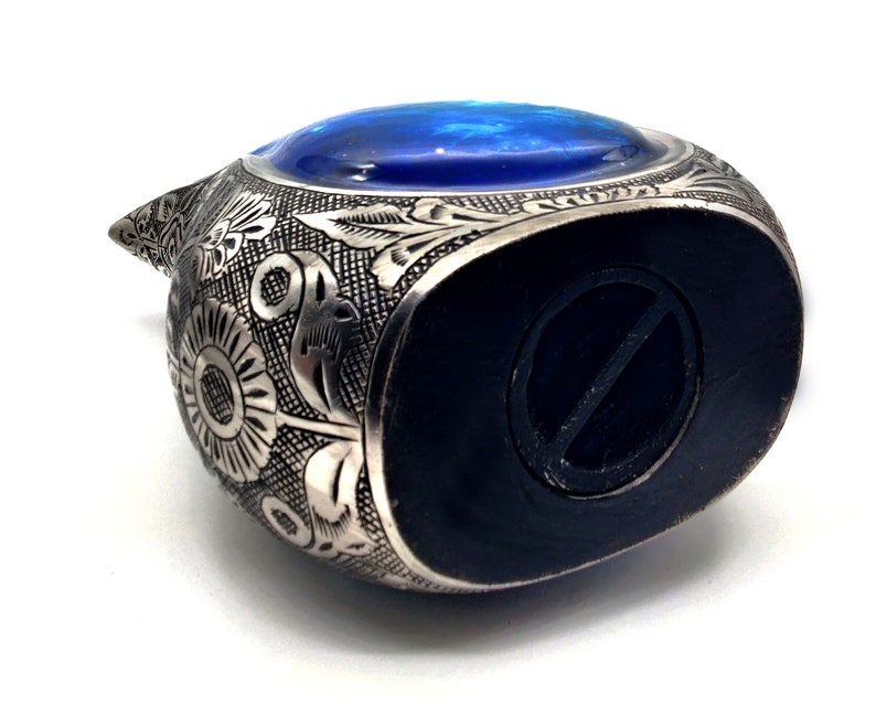Op de afbeelding: Een zilveren metalen spaarpot in de vorm van een vogel met een blauw glazen oog. De spaarpot heeft een ingewikkeld bloemmotief en een gleuf aan de onderkant voor munten.