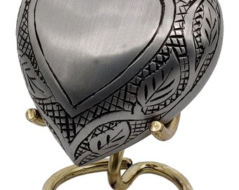 Pode incluir: Uma urna de cremação em forma de coração de prata com um design floral gravado. A urna está em um suporte de metal dourado.