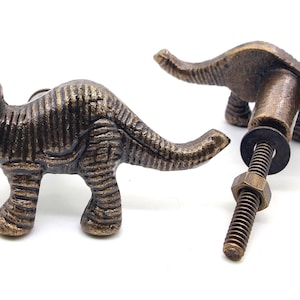 May include: Two bronze-colored dinosaur cabinet knobs. One knob is fully assembled, showing the screw and nut. The other knob is a standalone dinosaur. The knobs are detailed with textured skin and three horns.