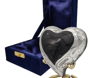 Pode incluir: Urna de cremação em forma de coração de prata com um design gravado. A urna está em um suporte dourado e vem em uma caixa de veludo azul.