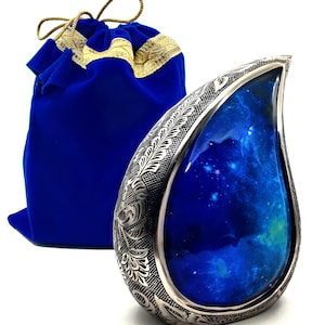 Peut inclure: Une urne funéraire en argent en forme de larme avec un motif floral gravé et un intérieur bleu sur le thème de la galaxie. L'urne est livrée avec un sac en velours bleu avec des garnitures dorées.