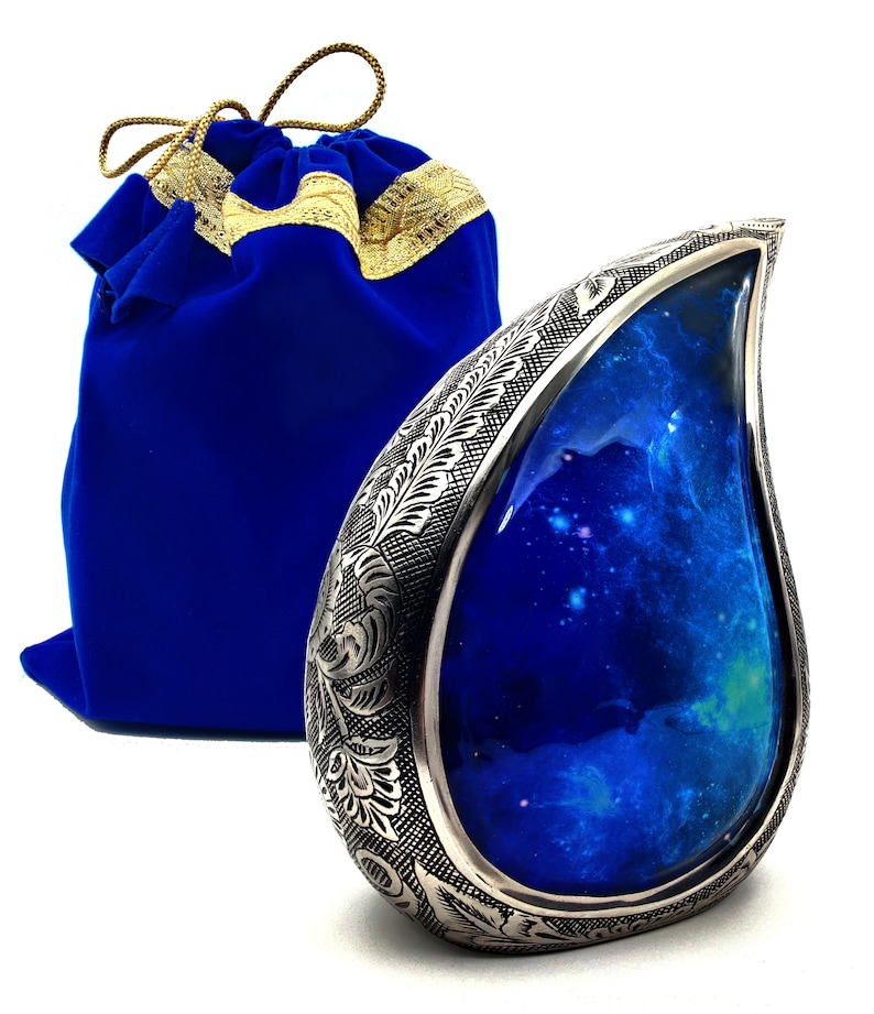 Op de afbeelding: Een zilveren urn in de vorm van een traan met een gegraveerd bloemenmotief en een blauw interieur met een galaxie-thema. De urn wordt geleverd met een blauwe fluwelen zak met gouden versiering.