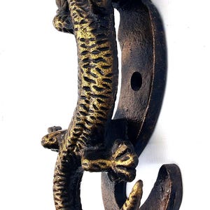 Puede incluir: Un gancho de pared de color bronce con un diseño detallado de lagarto. El gancho tiene una forma curva con un acabado texturizado y un tono dorado en el cuerpo del lagarto. El gancho está montado en una placa trasera curva.