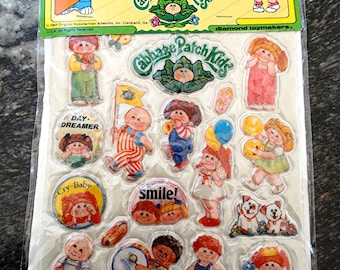 Puede incluir: Una hoja de pegatinas hinchadas con personajes de Cabbage Patch Kids. Las pegatinas son coloridas y presentan varios personajes, incluyendo un bebé tocando la batería, un bebé con un carro rojo y un bebé con un perro. Las pegatinas están empaquetadas en una bolsa de plástico transparente con un respaldo de cartón. El empaque incluye el texto "Puffy Stick-Ons" y "Cabbage Patch Kids".