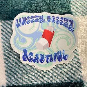 Puede incluir: Una pegatina blanca con un diseño de remolino azul y verde. La pegatina tiene el texto "Wheezy. Breezy. Beautiful" en letras azules y un gráfico de inhalador rojo y blanco.