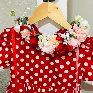 May include: A red polka dot dress with white spots, short sleeves, and a floral collar. The dress is hanging on a wooden hanger. The collar is decorated with pink, white, and red flowers. The dress has a white lace trim.