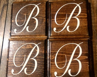 Puede incluir: Cuatro posavasos cuadrados de madera con tinte marrón oscuro. Cada posavasos presenta una gran letra "B" blanca en cursiva en el centro. Los posavasos están dispuestos en una formación cuadrada, mostrando el diseño de la letra.