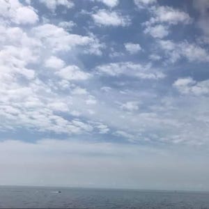 May include: A picturesque view of a blue sky dotted with soft, white clouds. The clouds are spread across the sky, giving it a textured look. Beneath the sky, a tranquil expanse of water extends to the horizon.