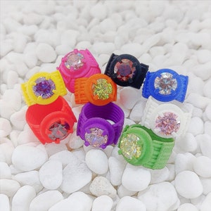 Puede incluir: Surtido de anillos de silicona de colores con piedras preciosas de diferentes colores. Cada anillo presenta una piedra preciosa de color diferente, incluyendo rojo, azul, morado, verde, amarillo y rosa.