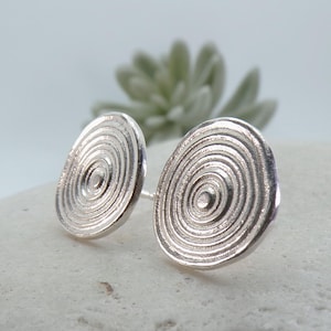 Puede incluir: Un par de pendientes de botón circulares de plata con un patrón de círculos concéntricos que irradian desde un punto central. La superficie metálica tiene un acabado pulido con una textura sutil, creando anillos en relieve en cada pendiente.