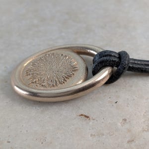 Könnte beinhalten: Ein goldfarbener Anhänger mit einem strukturierten, kreisförmigen Design, befestigt an einer schwarzen Lederschnur. Der Anhänger hat eine ovale Form mit einem erhabenen, sonnenstrahlenartigen Muster in der Mitte. Die Schnur ist geknotet.
