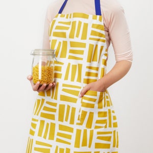 May include: A yellow and white patterned apron with a large pocket. The apron has a blue strap and is being worn by someone holding a glass jar filled with pasta.