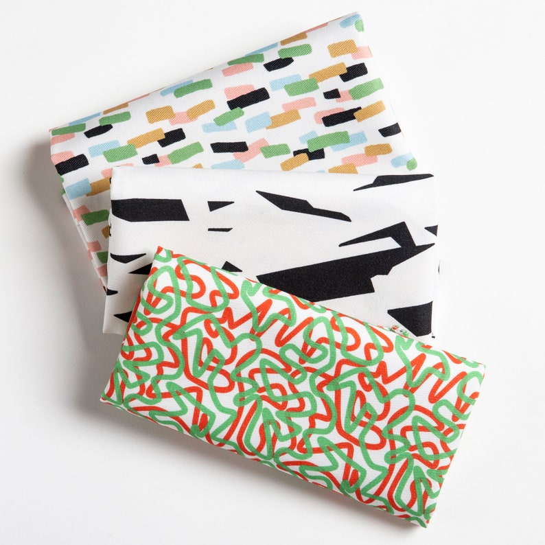 May include: Three fabric swatches with different geometric patterns. The top swatch has a white background with colorful abstract shapes. The middle swatch has a white background with black abstract shapes. The bottom swatch has a white background with green and red abstract shapes.