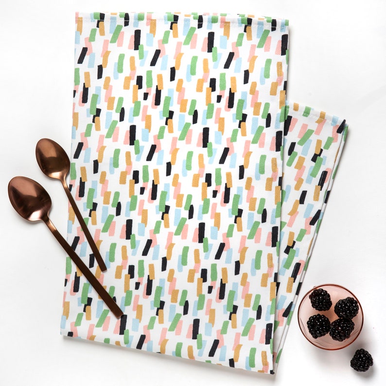 May include: A white kitchen towel with a colorful abstract pattern of black, green, blue, pink, and orange brushstrokes. The towel is folded and lying on a white surface.