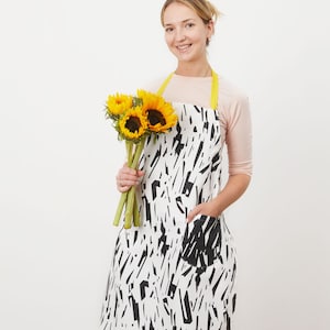 May include: A black and white patterned apron with a yellow strap. The apron has a large pocket on the front. A person is wearing the apron and holding a bunch of sunflowers.