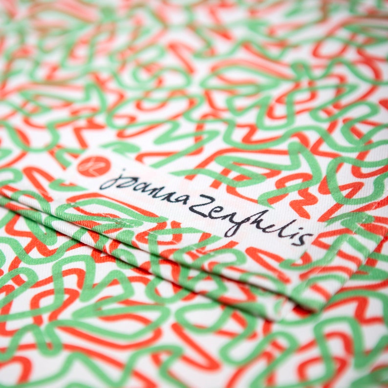 May include: White fabric with a repeating pattern of red and green squiggly lines. The fabric is folded and a label with the text "janna zephyrelis" is visible.