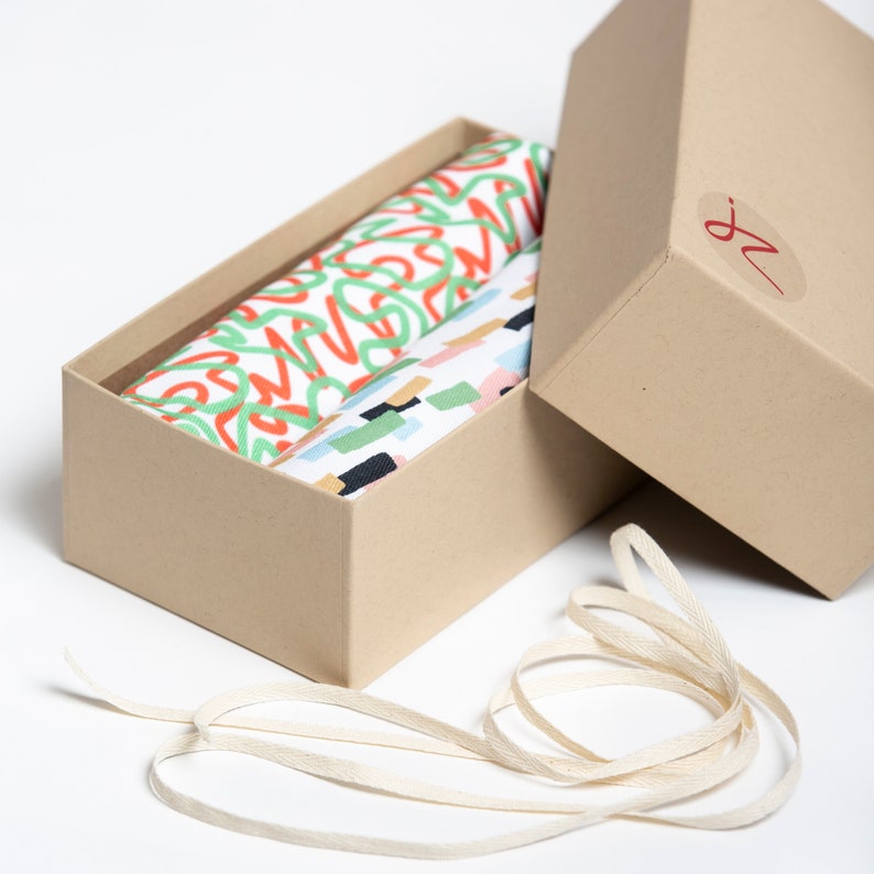 May include: A set of two fabric tea towels in a brown cardboard box with a white ribbon. One towel has a green and orange abstract pattern, the other has a colorful geometric pattern.