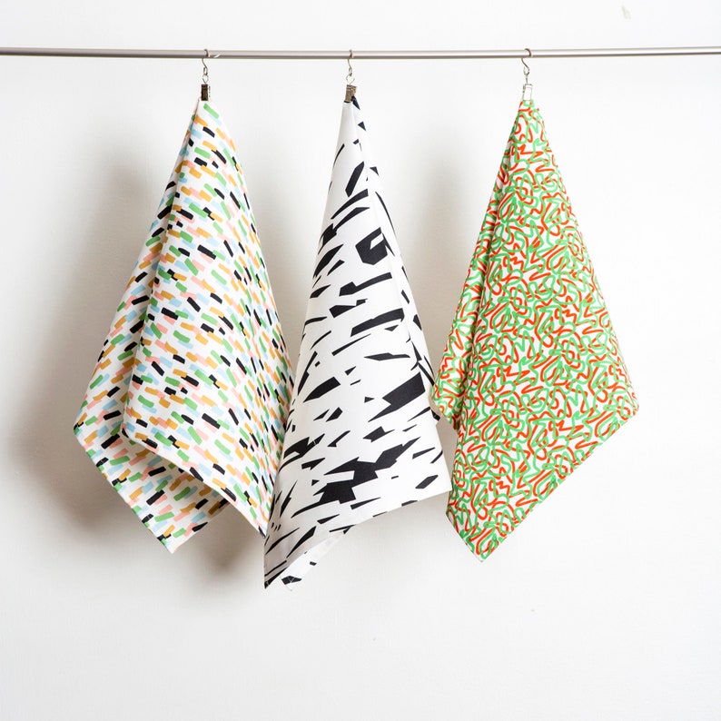 May include: Three kitchen towels hanging on a clothesline. The towels are white with different colorful patterns. The first towel has a pattern of colorful lines and dots. The second towel has a black and white pattern of abstract shapes. The third towel has a green and orange pattern of abstract shapes.