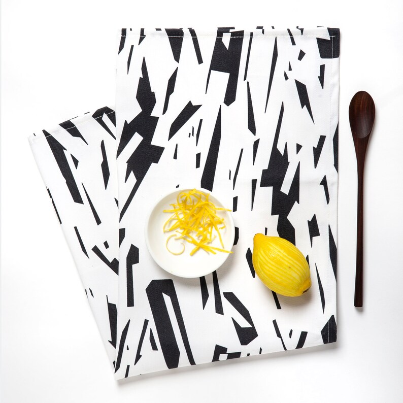May include: A white kitchen towel with a black geometric pattern. The towel is folded in half and has a small white bowl with lemon zest and a yellow lemon on top.