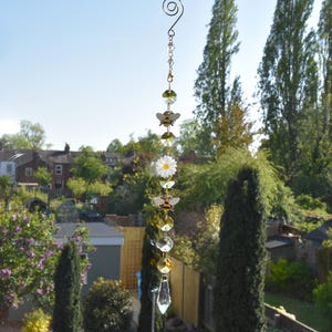 May include: A hanging suncatcher with a spiral top, incorporating crystal beads, faceted green and clear crystals, and bee and daisy charms. The suncatcher is suspended in front of a sunny outdoor scene with trees and houses.
