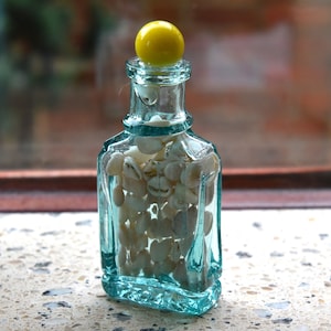 May include: A small, clear, turquoise glass bottle filled with small, white seashells. The bottle has a yellow, spherical stopper. The bottle is on a speckled surface.
