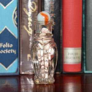 May include: A clear glass bottle with a fluted body filled with small, light-coloured seashells. The bottle is topped with a white marble featuring a prominent orange swirl pattern. The glass container has a narrow neck and a textured, decorative base.