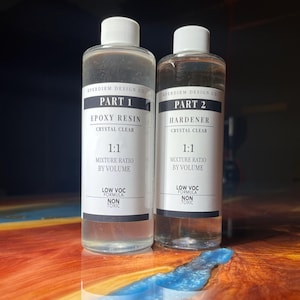 Puede incluir: Dos botellas transparentes de resina epoxi y endurecedor, etiquetadas como "Parte 1" y "Parte 2" con texto "Cristalino". Las botellas tienen tapas blancas y etiquetas con texto negro. Se ve una pequeña cantidad de resina azul.
