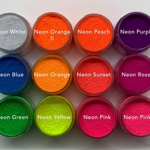 Op de afbeelding: Twaalf ronde potjes met levendige pigmentpoeders in diverse neonkleuren, waaronder wit, oranje, perzik, paars, blauw, sunset, rose, groen, geel en roze. Elk potje is voorzien van een label met de bijbehorende kleurnaam in witte tekst.