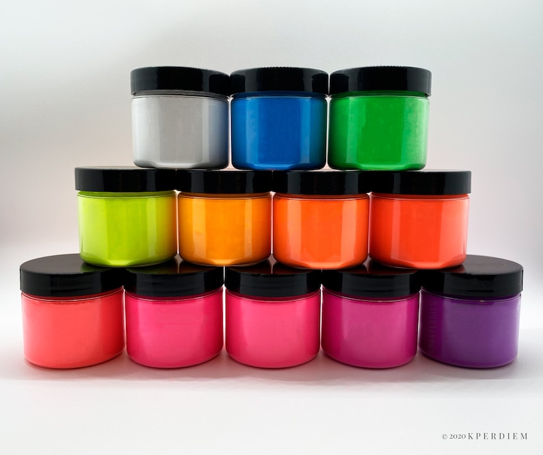 Op de afbeelding: Een verzameling van twaalf cilindrische potten met zwarte deksels, gerangschikt in een piramidevorm. De potten bevatten levendige verven in zilver, blauw, groen, geel, oranje, rood, roze, magenta en paars. De tekst 2020 KPERDIEM is zichtbaar.