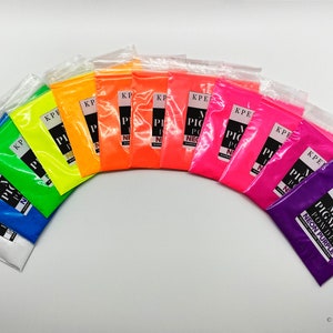 May include: A set of 12 small plastic bags containing neon pigment powder in various colors, including white, blue, green, yellow, orange, red, pink, and purple. The bags are arranged in a semi-circle on a white surface. Each bag has a label that reads "KPERDIEM MICA PIGMENT POWDER NEON [COLOR]"
