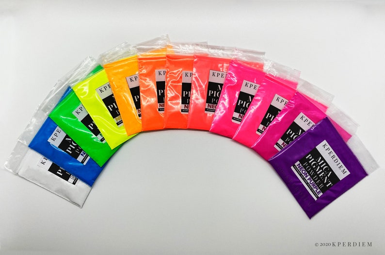 May include: A set of 12 small plastic bags containing neon pigment powder in various colors, including white, blue, green, yellow, orange, red, pink, and purple. The bags are arranged in a semi-circle on a white surface. Each bag has a label that reads "KPERDIEM MICA PIGMENT POWDER NEON [COLOR]"