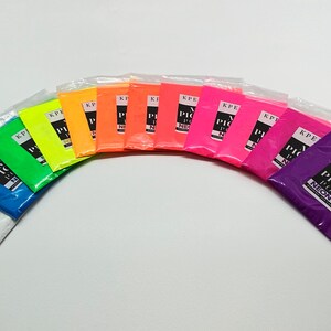 May include: A set of 12 small bags of neon mica pigment powder. The bags are arranged in a semi-circle, with the colors ranging from white to purple. Each bag has a black label with the text "KPERDIEM MICA PIGMENT POWDER NEON [COLOR]" printed on it.