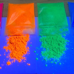 Op de afbeelding: Vier doorzichtige plastic zakjes met fijn fluorescerend pigmentpoeder in levendige tinten roze, oranje, groen en blauw. Elk zakje is aan de onderkant gedeeltelijk geopend, waardoor een klein hoopje van het losse, gekleurde poeder naar buiten komt.