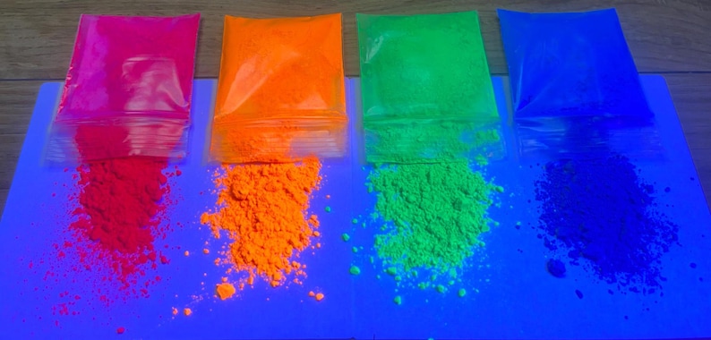 Op de afbeelding: Vier doorzichtige plastic zakjes met fijn fluorescerend pigmentpoeder in levendige tinten roze, oranje, groen en blauw. Elk zakje is aan de onderkant gedeeltelijk geopend, waardoor een klein hoopje van het losse, gekleurde poeder naar buiten komt.