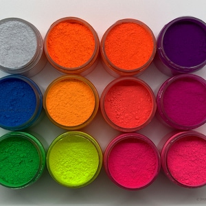 May include: Twelve small jars of neon pigment powder in various colors, including orange, pink, green, yellow, blue, and purple. The jars are arranged in a grid pattern on a white surface.