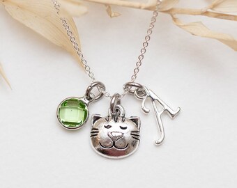 May include: A silver-toned necklace featuring three charms: a round light green faceted stone, a small cat face pendant with whiskers and closed eyes, and a silver letter A charm, all suspended from a delicate silver-toned chain.