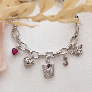May include: A silver charm bracelet featuring a selection of charms: a red heart, a heart, a perfume bottle, a bee, and the letter "J". The bracelet is presented on a white surface, with dried leaves and a pink ribbon in the background.
