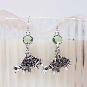 May include: Silver turtle earrings with green gemstone accents. The earrings are hanging from a white candle holder.