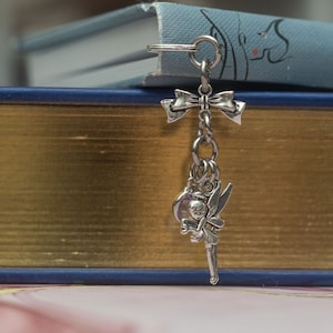 May include: Silver bookmark featuring a fairy with wings and a wand, holding a small round object. The bookmark is attached to a silver bow and chain.